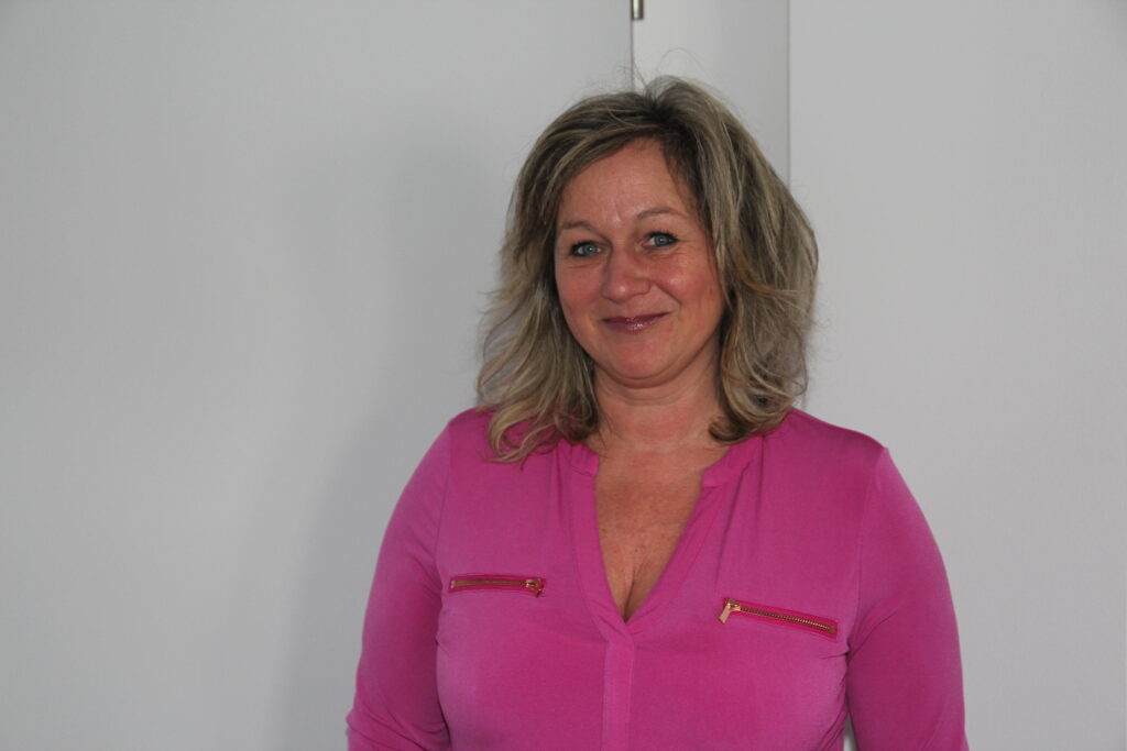 Medizinische Mitarbeiterin mit pinker Bluse vor neutralem Hintergrund.