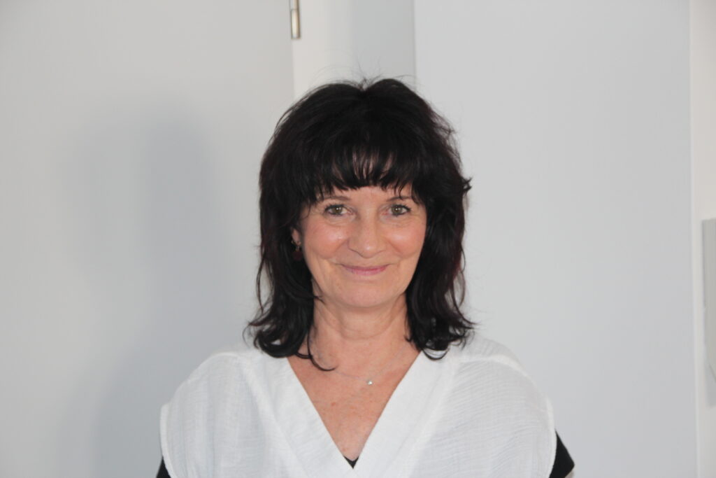 Medizinische Mitarbeiterin mit dunklen Haaren und weißer Bluse vor heller Wand.